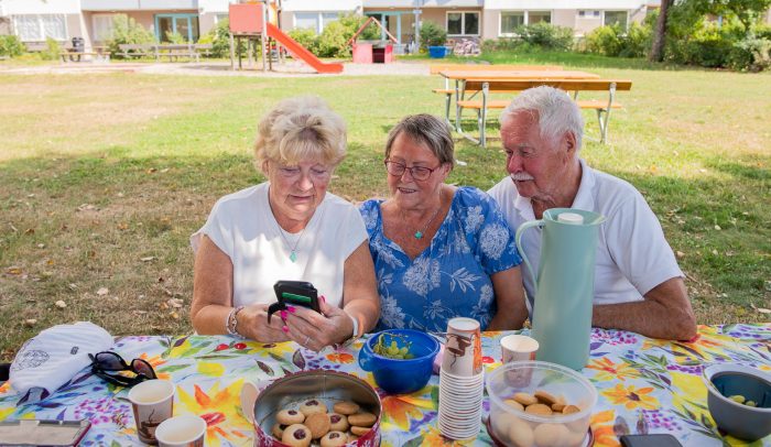 Tre äldre personer har samlats utomhus runt en mobiltelefon.